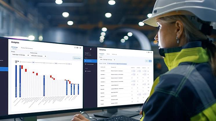 A woman wearing a hard hat and safety vest is focused on two computer screens in a work environment.