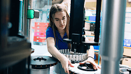 A woman works diligently on a factory machine, surrounded by tools and equipment in a busy industrial environment.