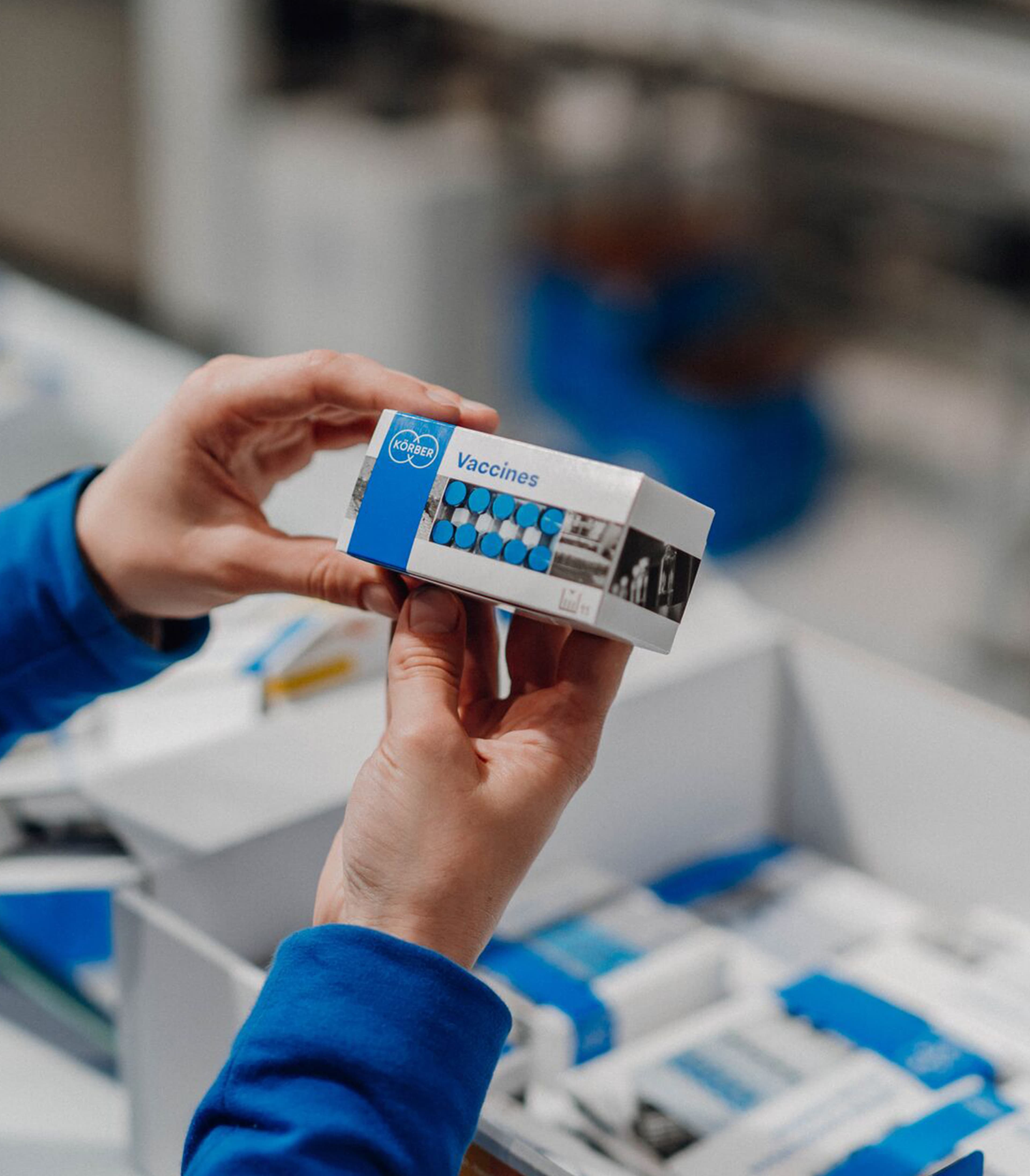 Hands holding a Körber vaccine packaging box inside a pharmaceutical production facility.