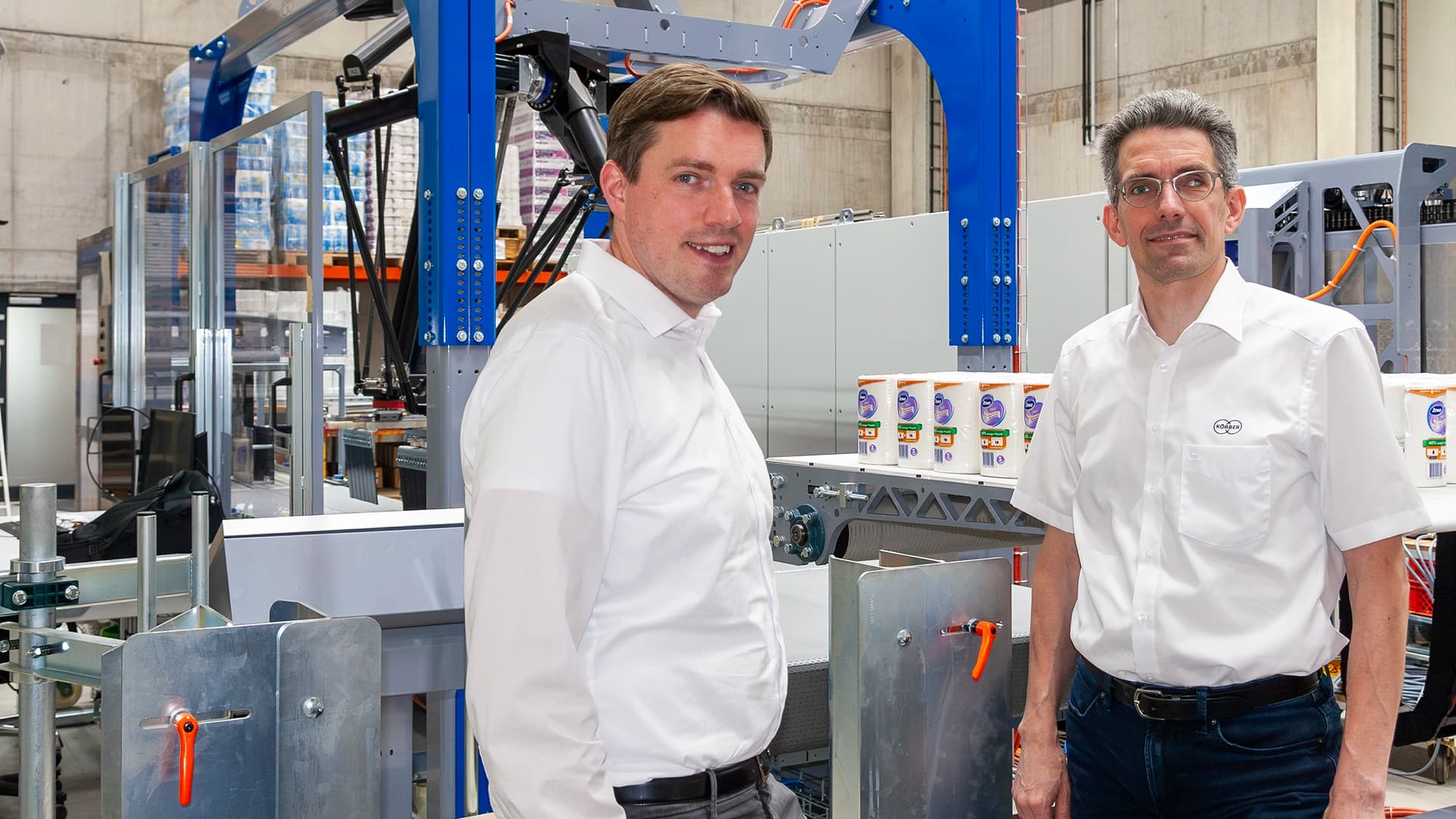Two men stand in front of a PA15 layer palletizer machine in a factory setting.