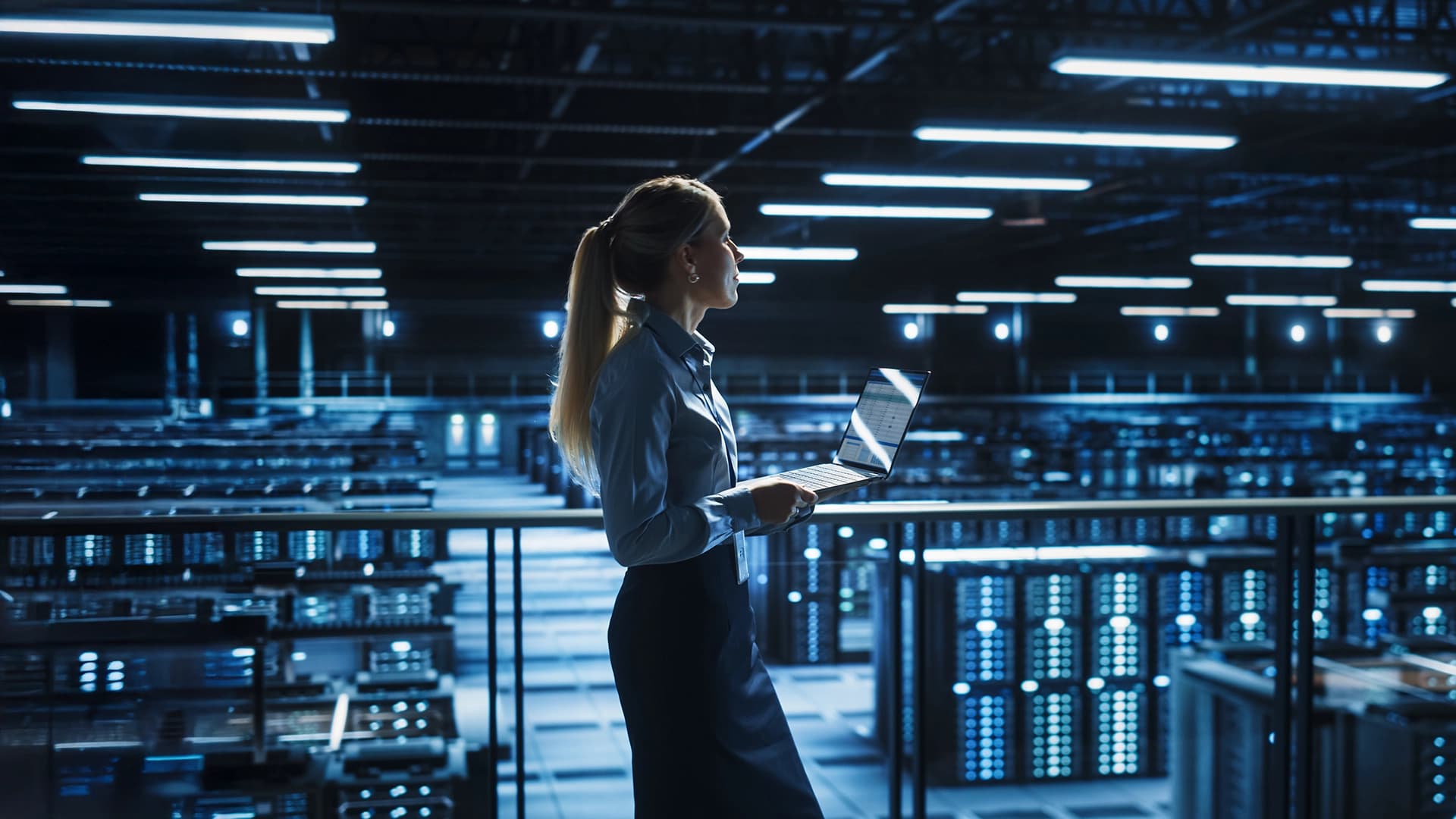 A woman stands amidst a vast server room, featuring multiple server racks and illuminated control panels