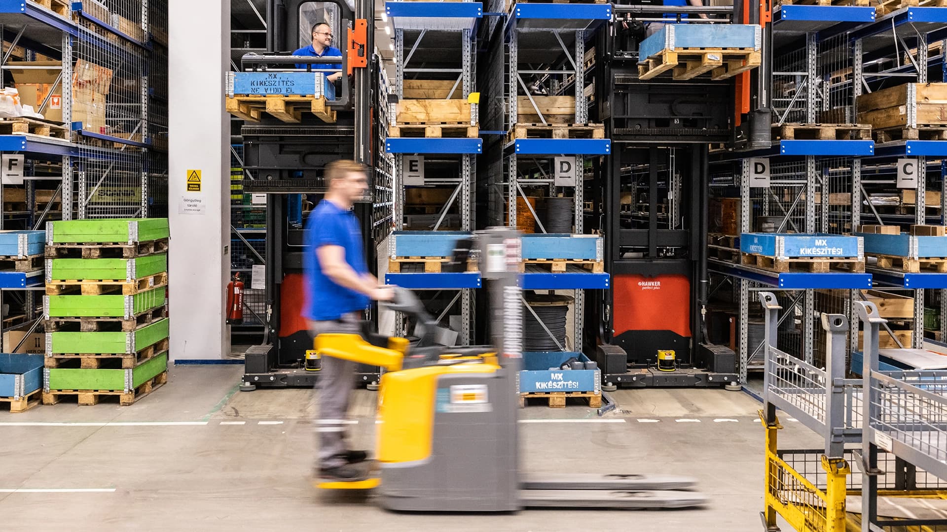 A man operates a forklift inside a warehouse, maneuvering between shelves filled with boxes.