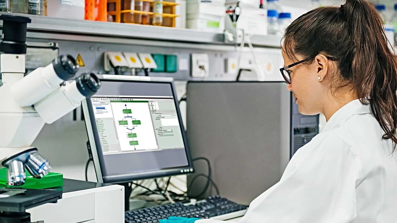 A woman wearing a lab coat is engaged in computer work, surrounded by laboratory equipment.