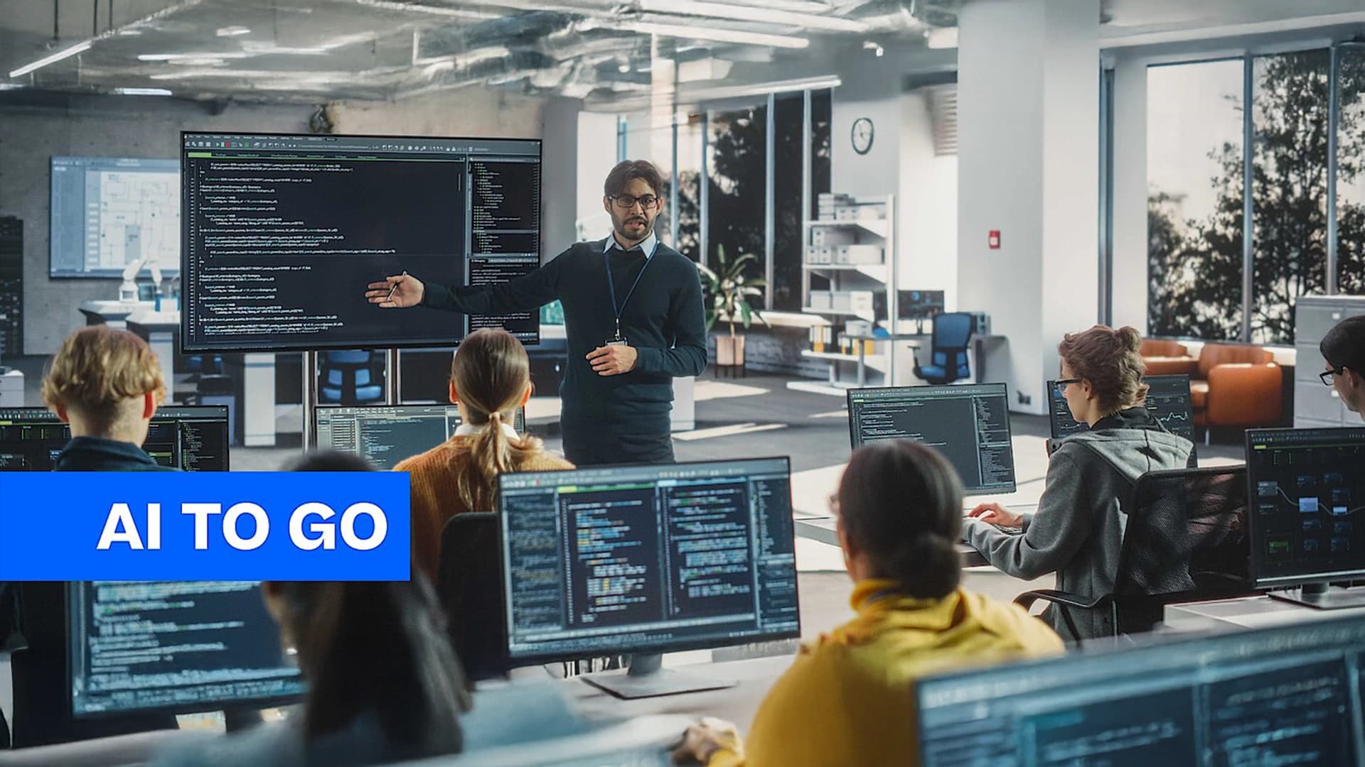 A group of people working at computers, while manager is standing up explaining them something.
