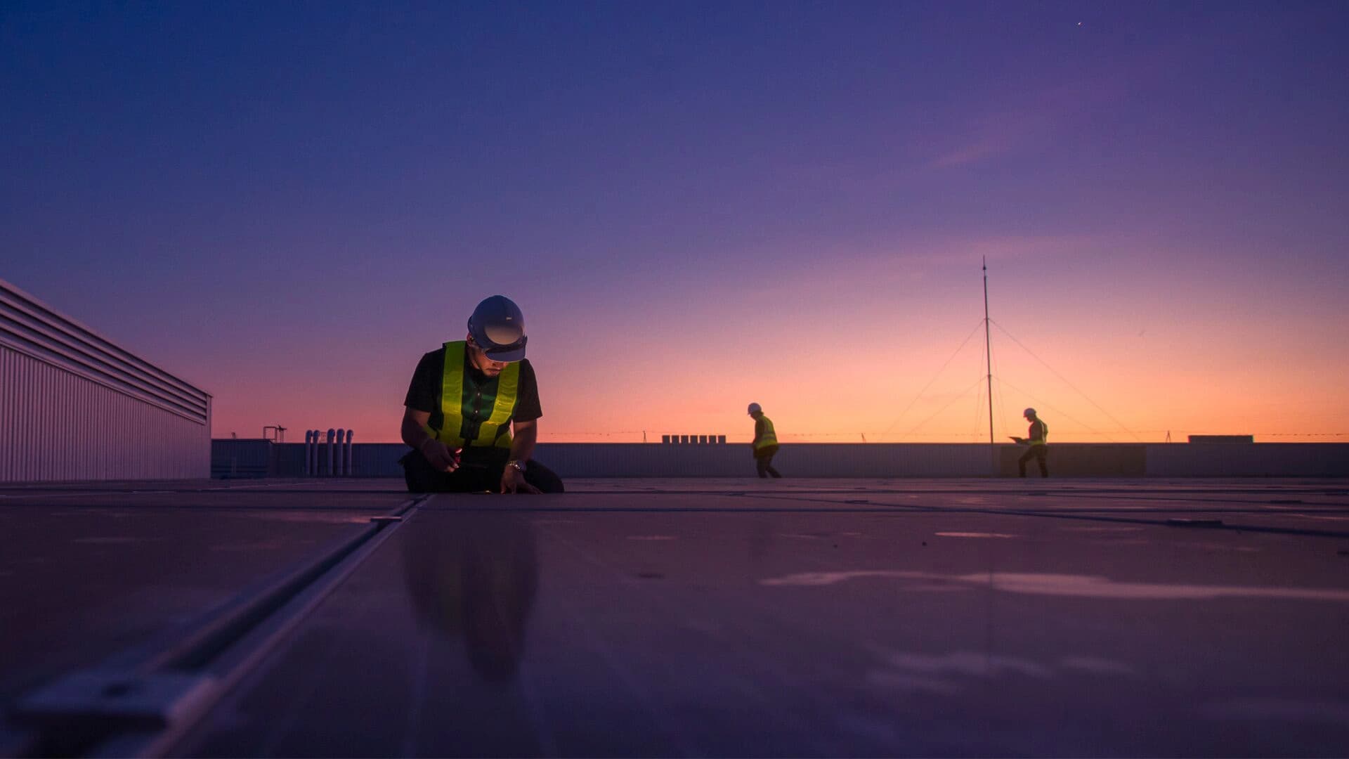Workers on a building roof silhouetted against a vibrant sunset sky.