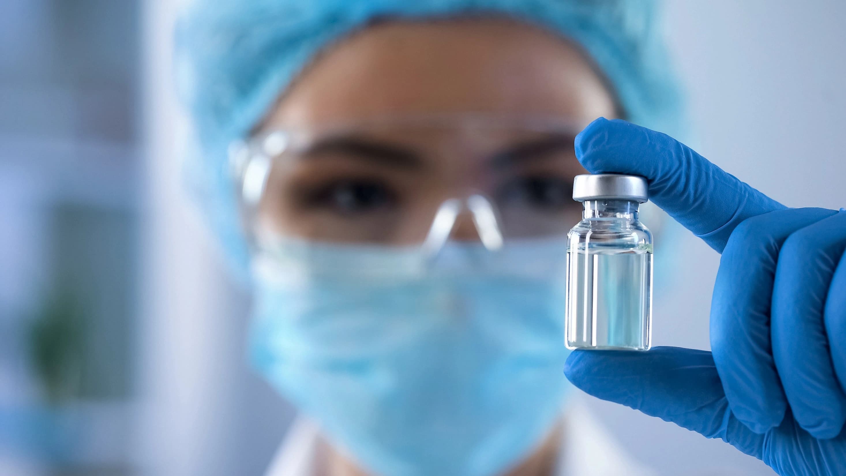 A woman wearing a blue surgical mask and gloves holds a vial, indicating a medical or laboratory setting.