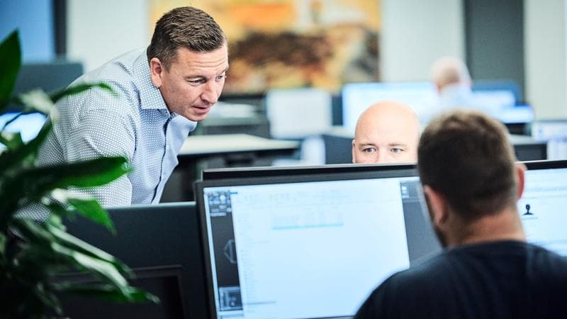 Two man working on computers while one of them supervises them.