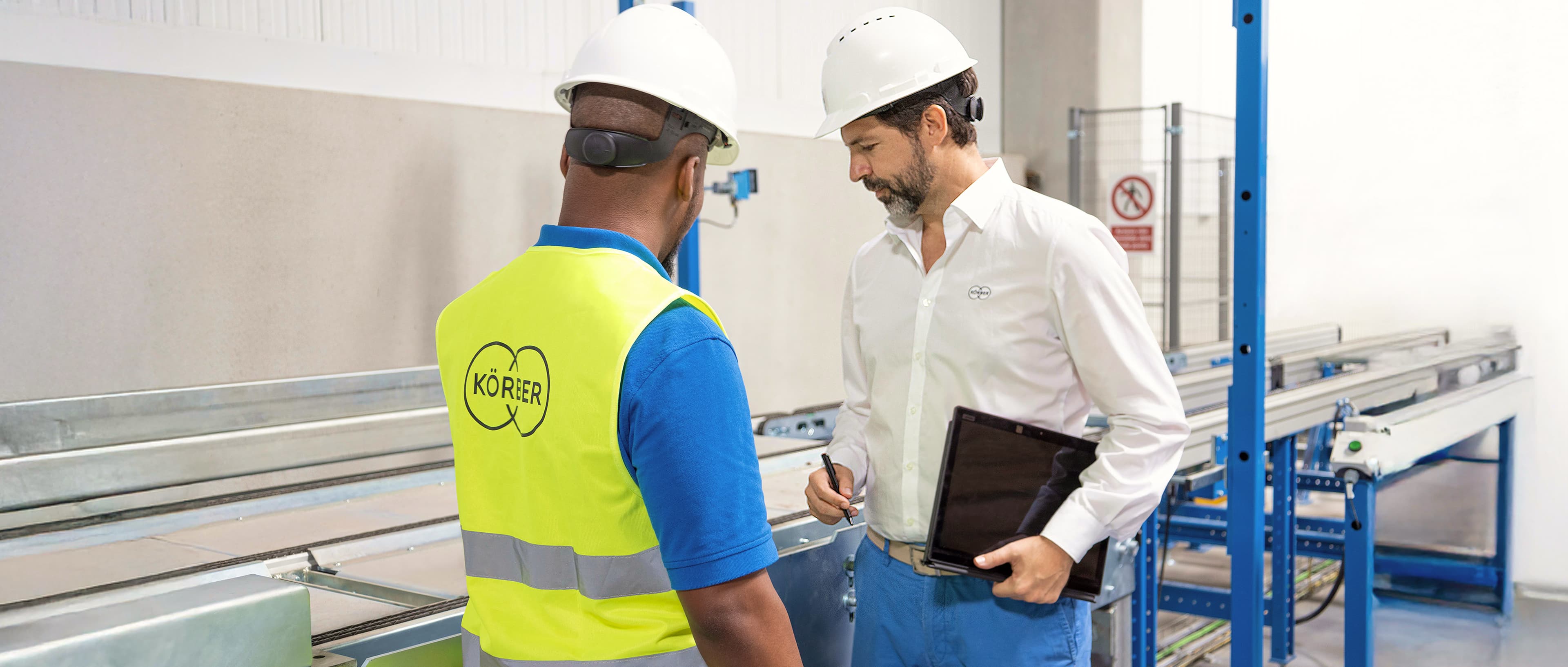 A Körber employee in a high visibility vest inspecting a machine with a Körber consultant in a white shirt
