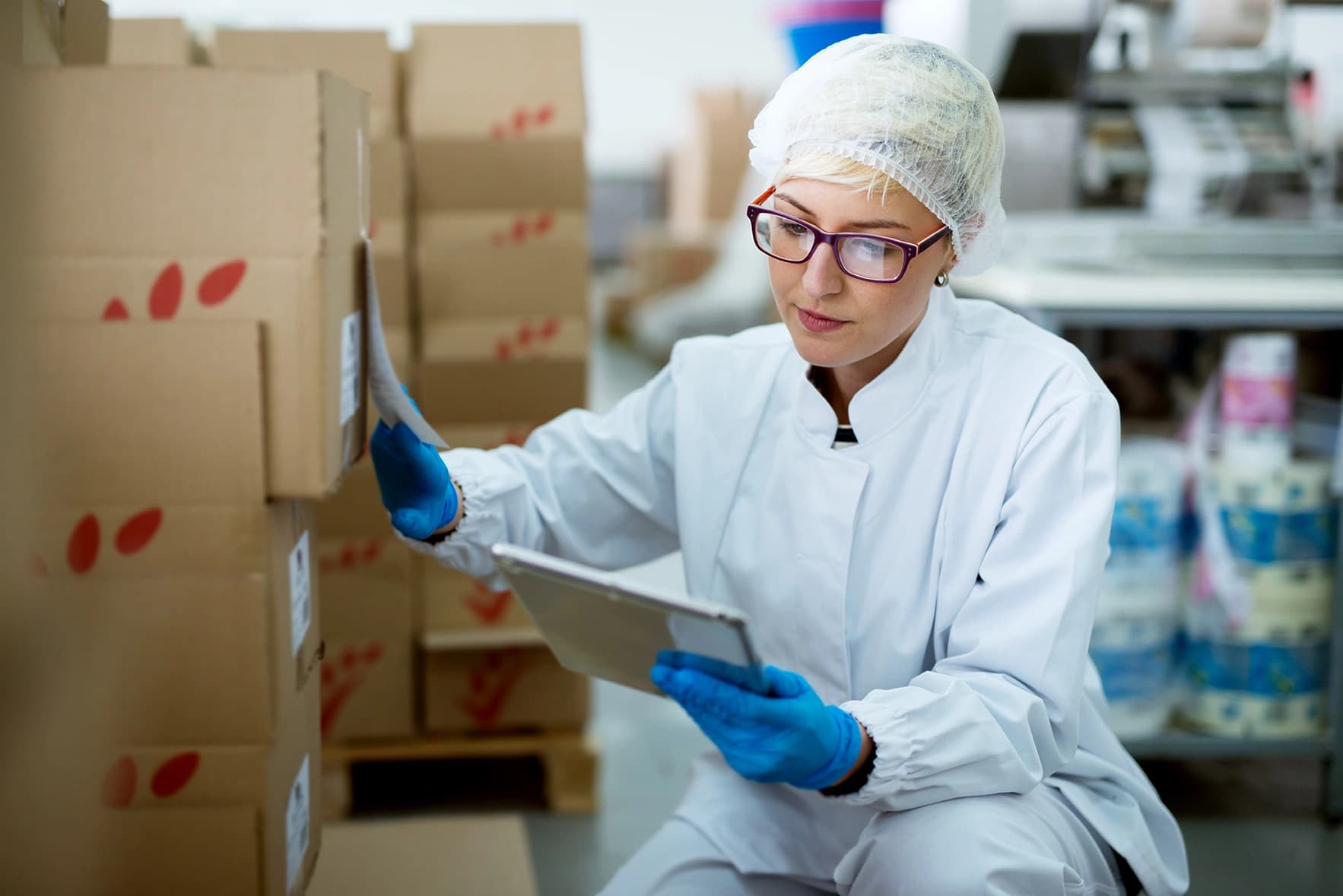 A woman in a lab coat is focused on her tablet, next to several carton boxes.