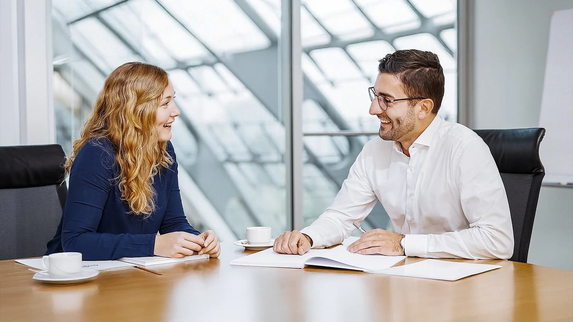 A man and woman are seated at a table in an office, discussing work-related topics.