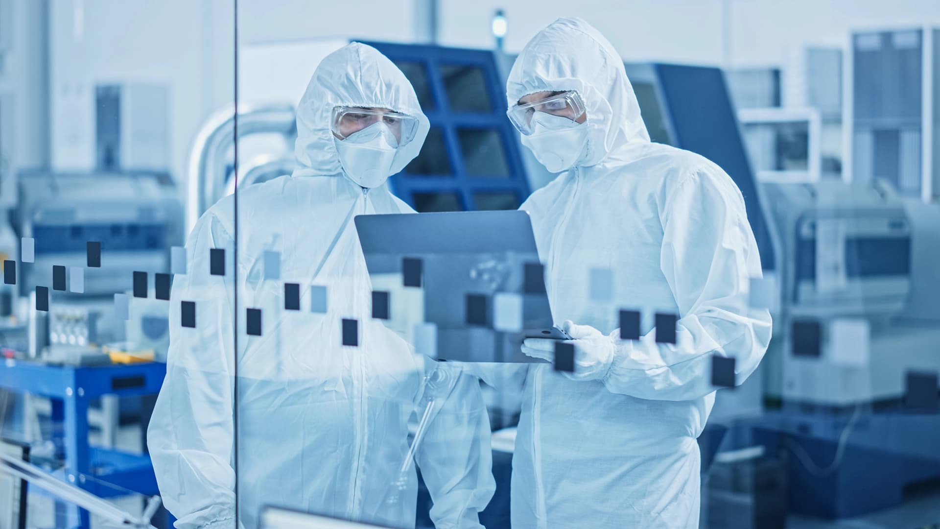 Two people in white protective suits analyze data on a laptop in a pharmaceutical production setting.